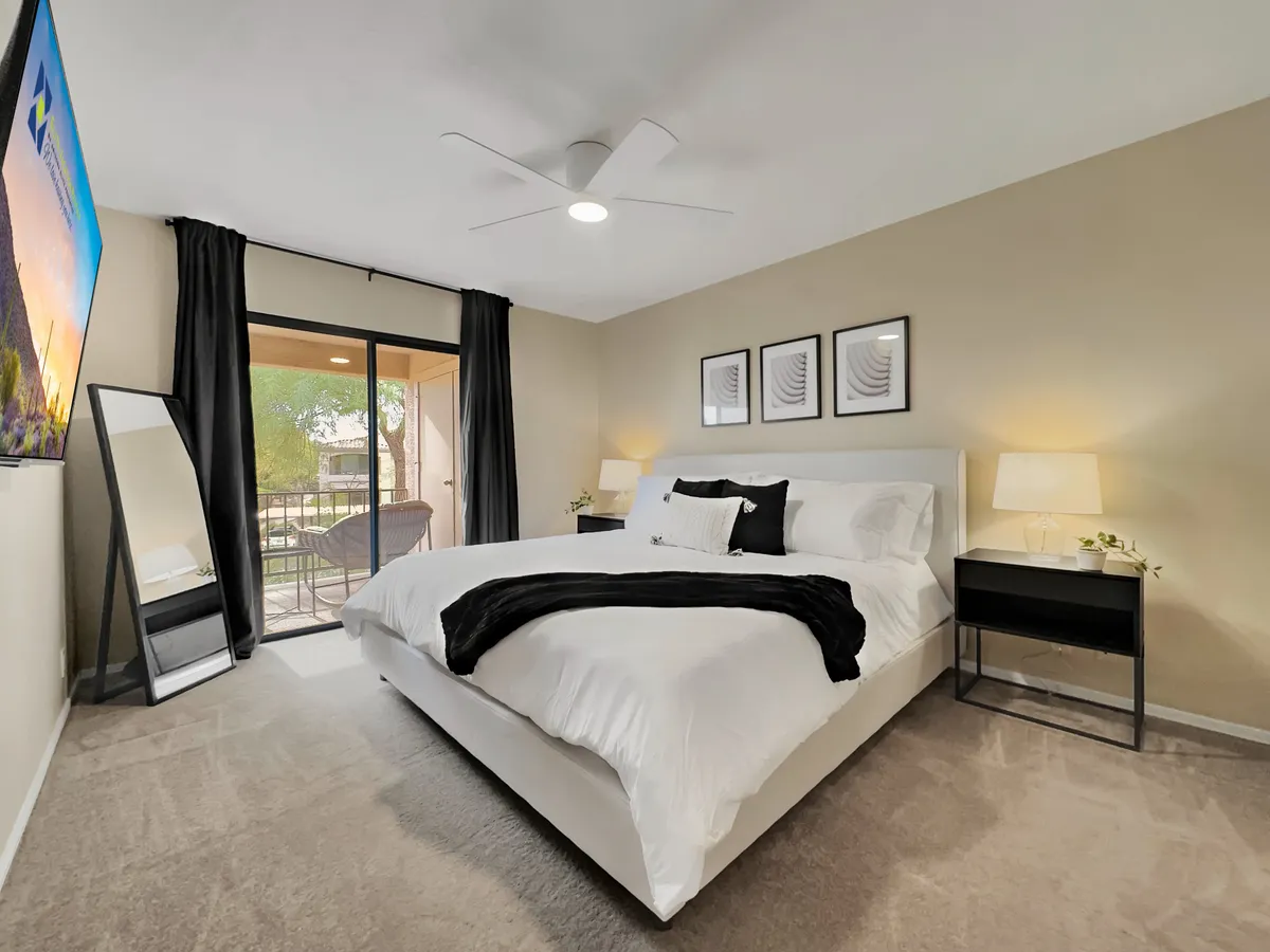 A modern king-size bedroom featuring crisp white bedding, a large mirror that enhances space, a balcony door with flowing curtains, minimalist artwork, and bedside lamps. The suite offers natural light and a sense of openness.