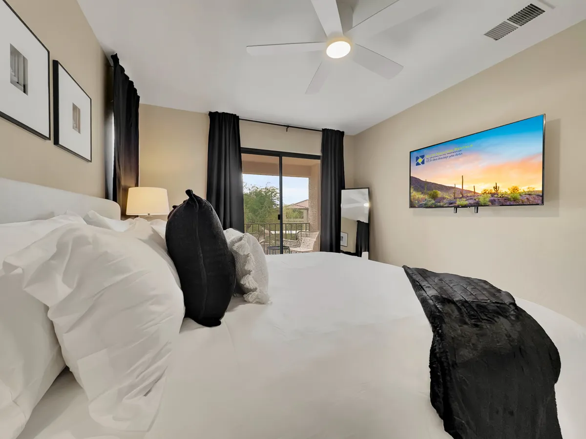 A contemporary bedroom with white linens, a large window overlooking the balcony, a TV mounted on the wall, black curtains, and decorative pillows. The space feels both comfortable and modern, with amenities for relaxation and entertainment.