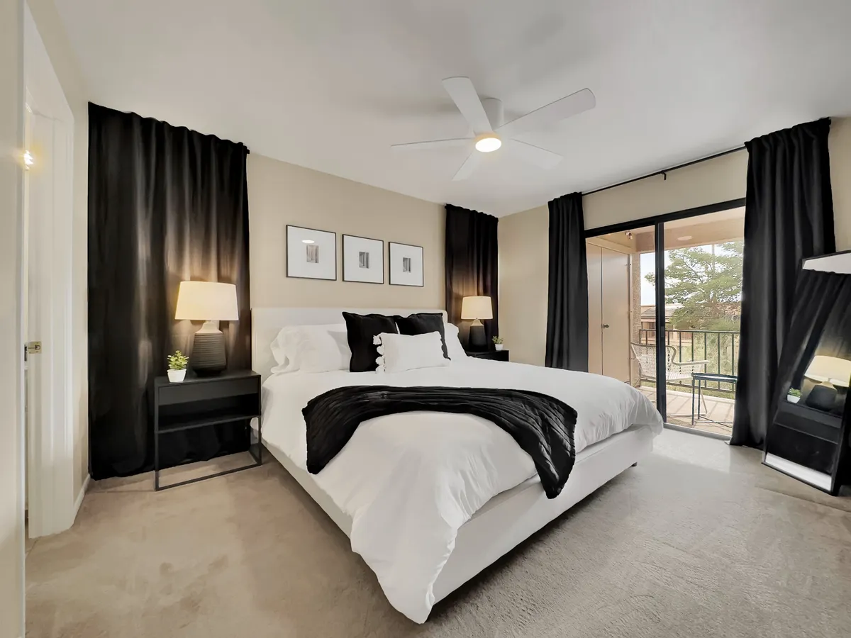 A serene bedroom setting with a bed adorned with decorative pillows, black curtains for privacy, nightstands with soft-lit lamps, framed artwork, and a glass door leading to a balcony. The space feels restful and private.