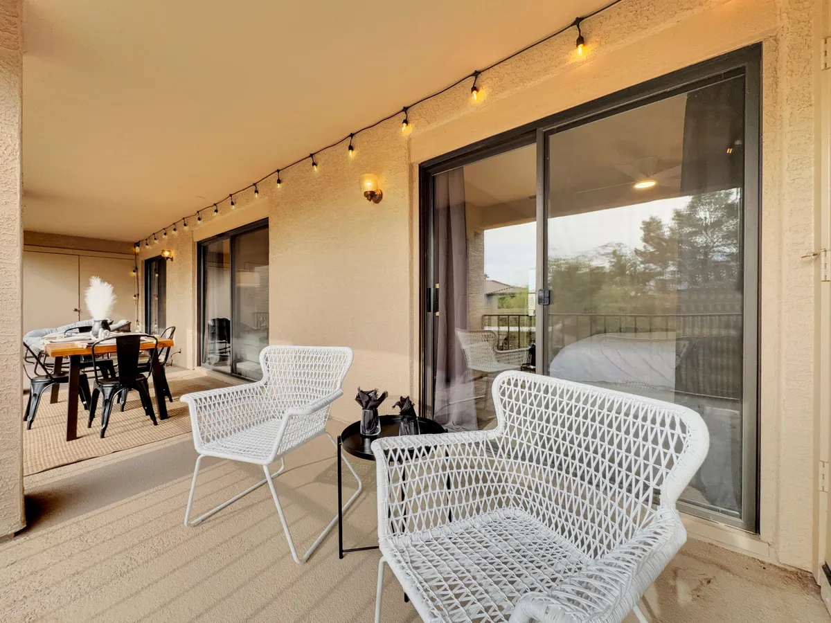An inviting outdoor patio set with white wicker chairs, a small black table, string lights overhead, and a dining table with chairs. The setting radiates a casual, communal atmosphere, perfect for outdoor meals or relaxed evenings with friends.