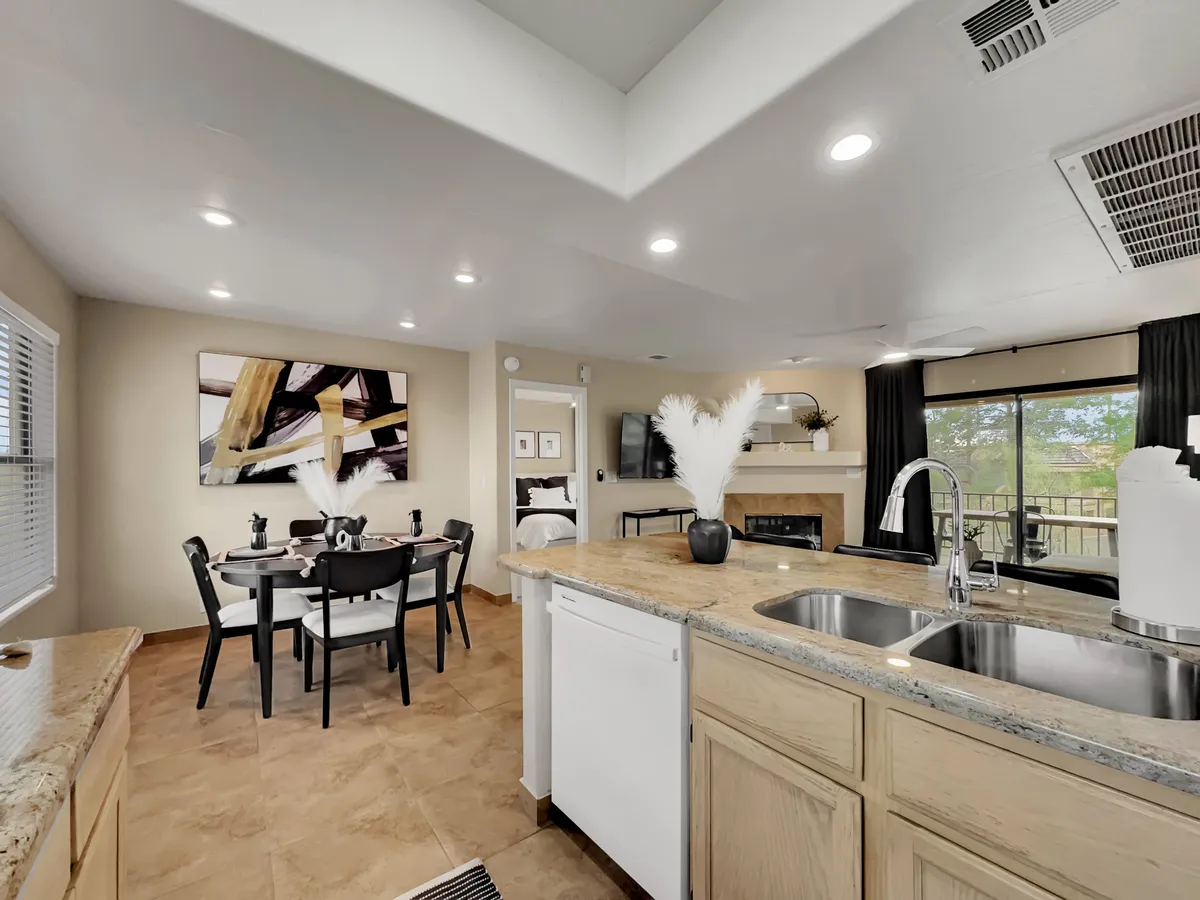 A kitchen island with a sink takes center stage, with a dining table nearby. The scene features inviting artwork, a fireplace, and large windows that offer an abundance of natural light and outdoor views.