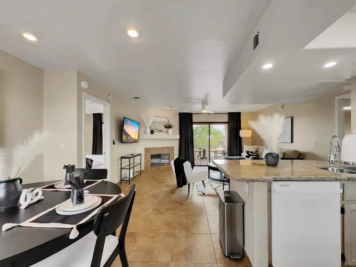 A cozy, inviting living area featuring a dining table, comfortable seating, a fireplace, large windows, and a television. The space is filled with natural light, and the windows offer glimpses of the balcony beyond, integrating the indoor and outdoor environments.