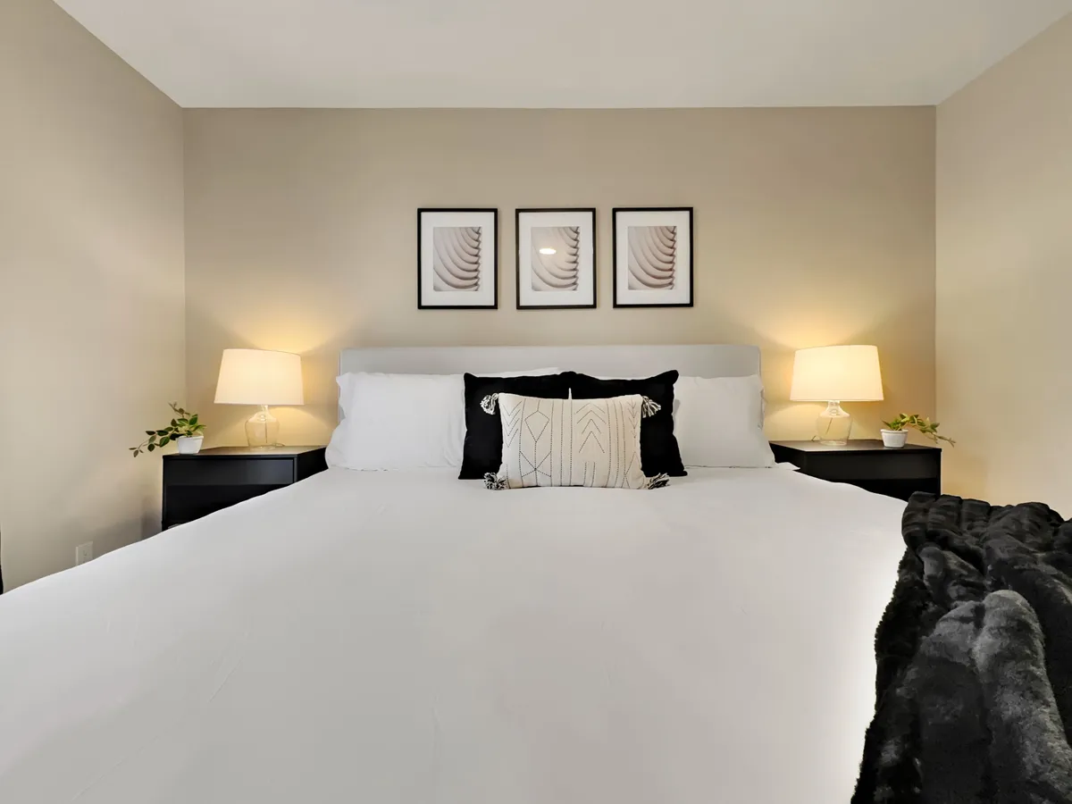 A tranquil master suite with a large king bed, plush decorative pillows, framed wall art, and matching nightstands with lamps. The serene color palette and soft lighting create a restful atmosphere ideal for relaxation and privacy.