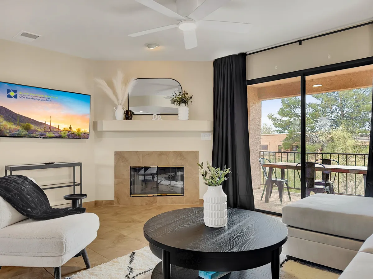 A comfortable living area with a prominent fireplace, mounted television, coffee table, and a direct view through glass doors to a balcony. The inviting seating and glowing fire complete the welcoming atmosphere.